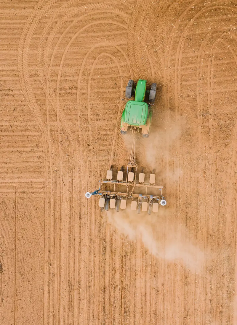 Overhead view of a green tractor pulling a seed drill across a dry, tilled field, raising dust.